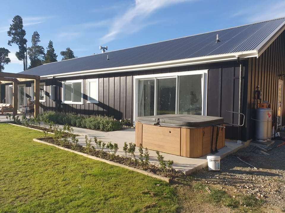 A single-storey house with black vertical board cladding and white window frames, a spa pool on the patio and a new lawn