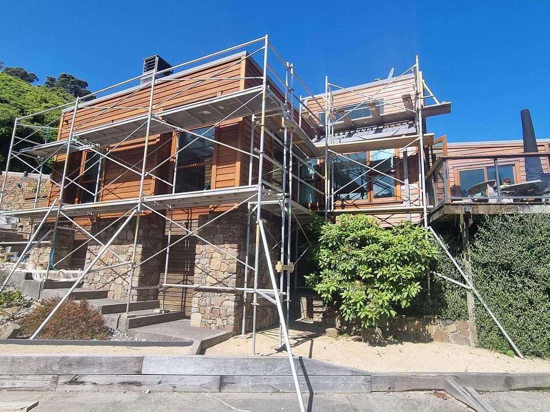 A two-storey house clad in cedar boards on a stone base, wrapped in scaffolding for exterior work