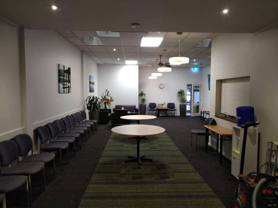 A long staff room with white walls, rows of chairs, round white tables and a green patterned carpet