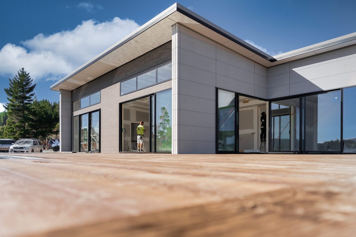 The corner of a modern house with pale grey cladding and floor-to-ceiling glass, seen from low across a timber deck, with a painter in a hi-vis shirt inside