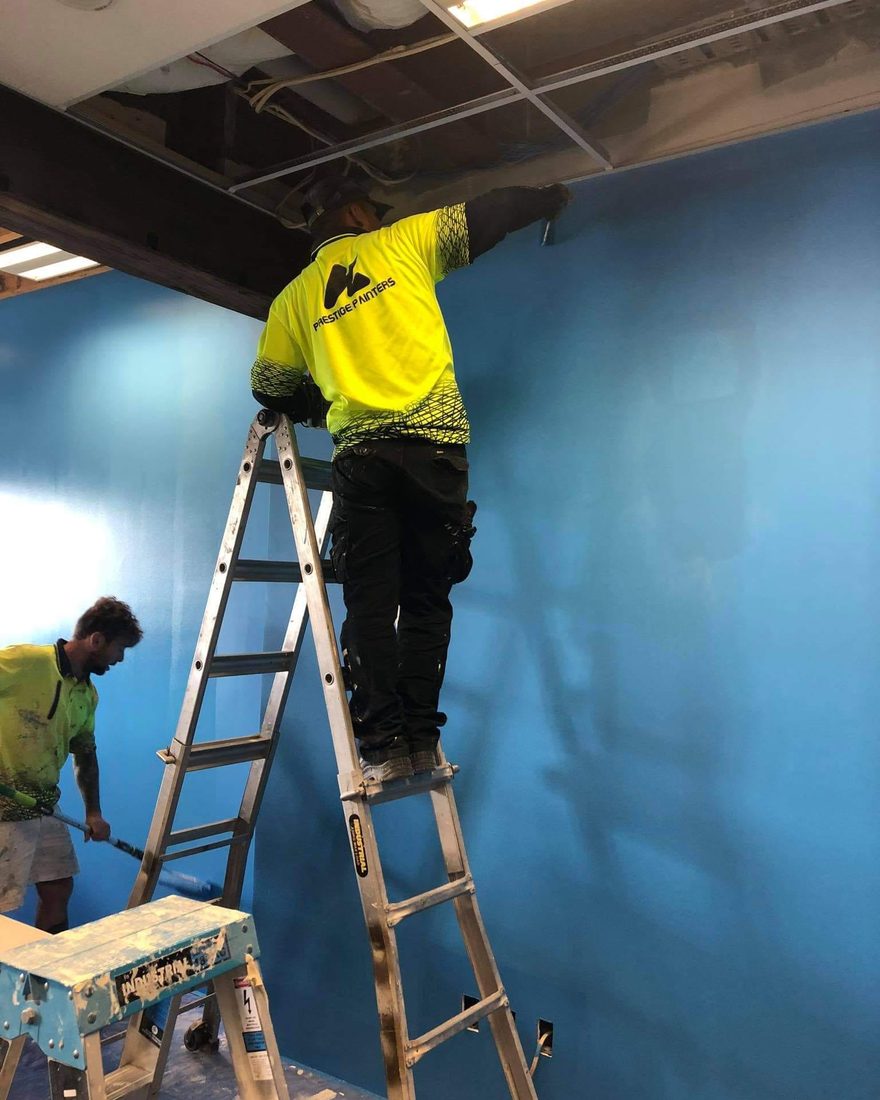Two painters in hi-vis shirts on a ladder and a trestle, painting a tall interior wall blue