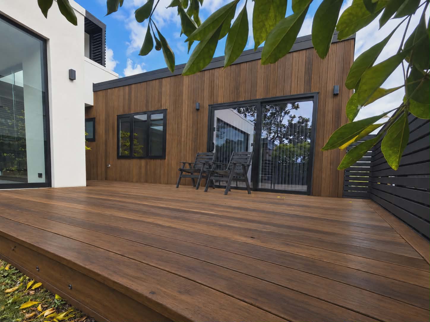 A timber deck in front of a wall of vertical stained cedar boards with black-framed windows and sliding doors, two outdoor chairs and leaves hanging over the foreground