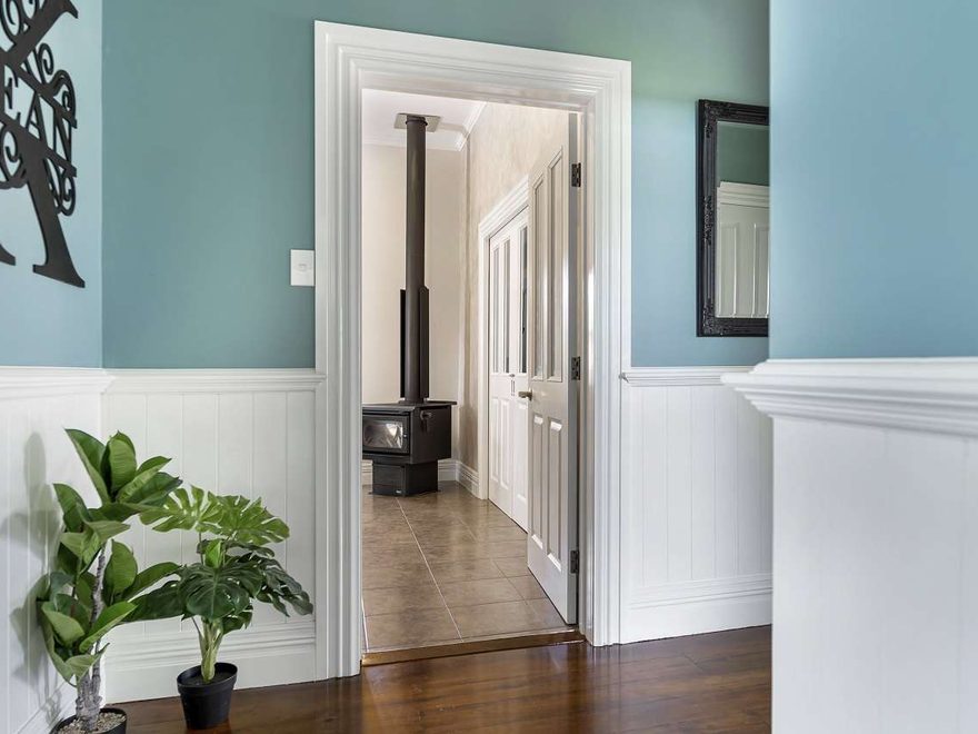 A hallway with blue-green walls above white panelling, looking through a white doorway to a room with a wood burner